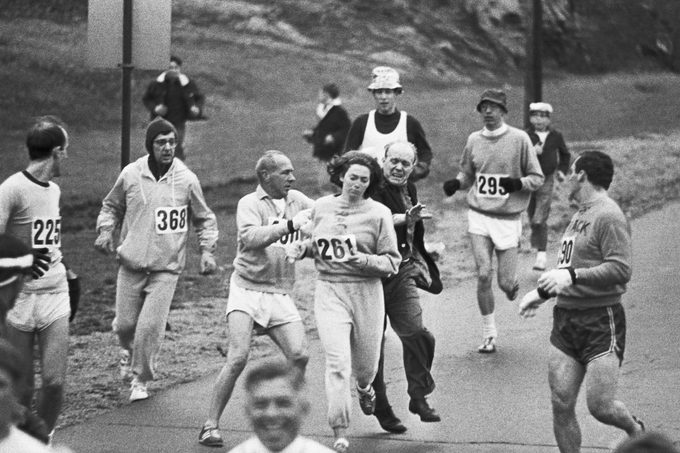 black and white photograph of Trainer Jock Semple -- in street clothes -- enters the field of runners (left) to try to pull Kathy Switzer (261) out of the race